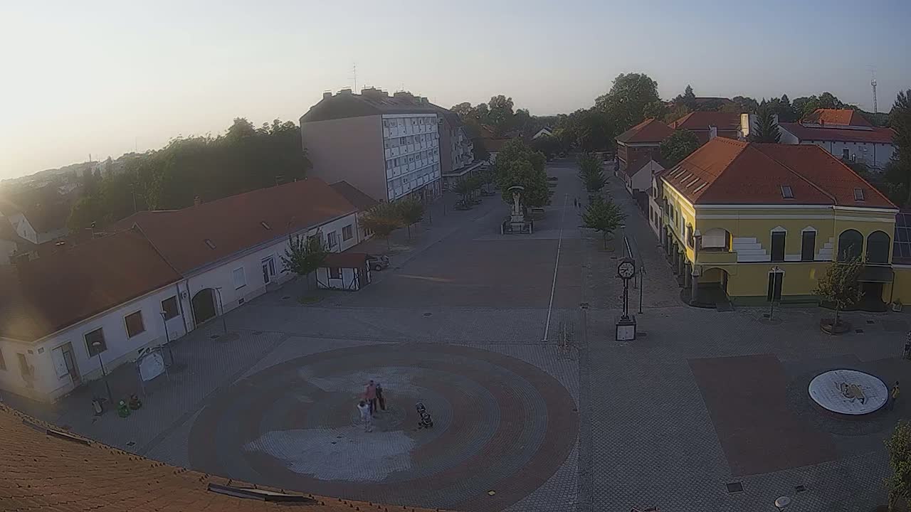 A high-angle, long shot of a town square on a sunny day shows several buildings surrounding a paved area with a circular fountain and a few people near it.
