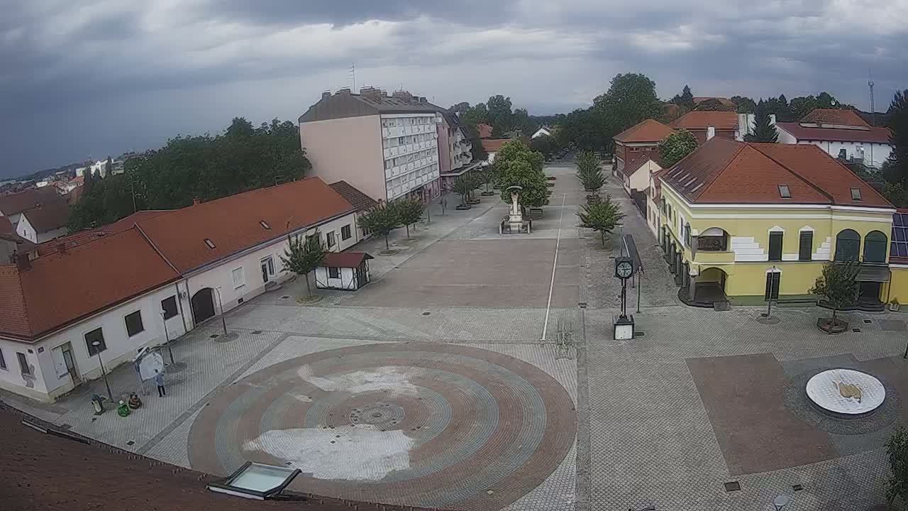 A town square with low buildings under a cloudy sky, featuring a circular paved area in the center and a monument.