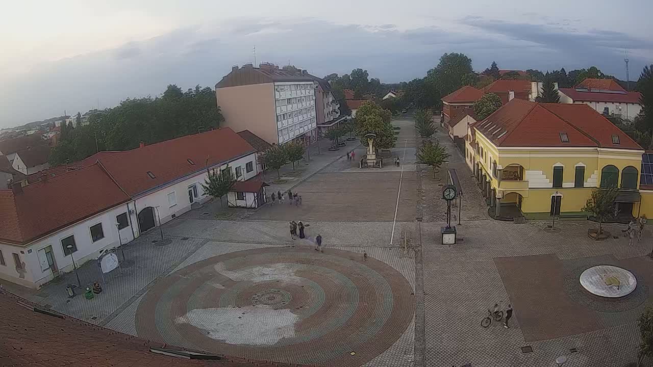 An overcast sky hangs over a town square with several low buildings, a central fountain, and a few people walking around.
