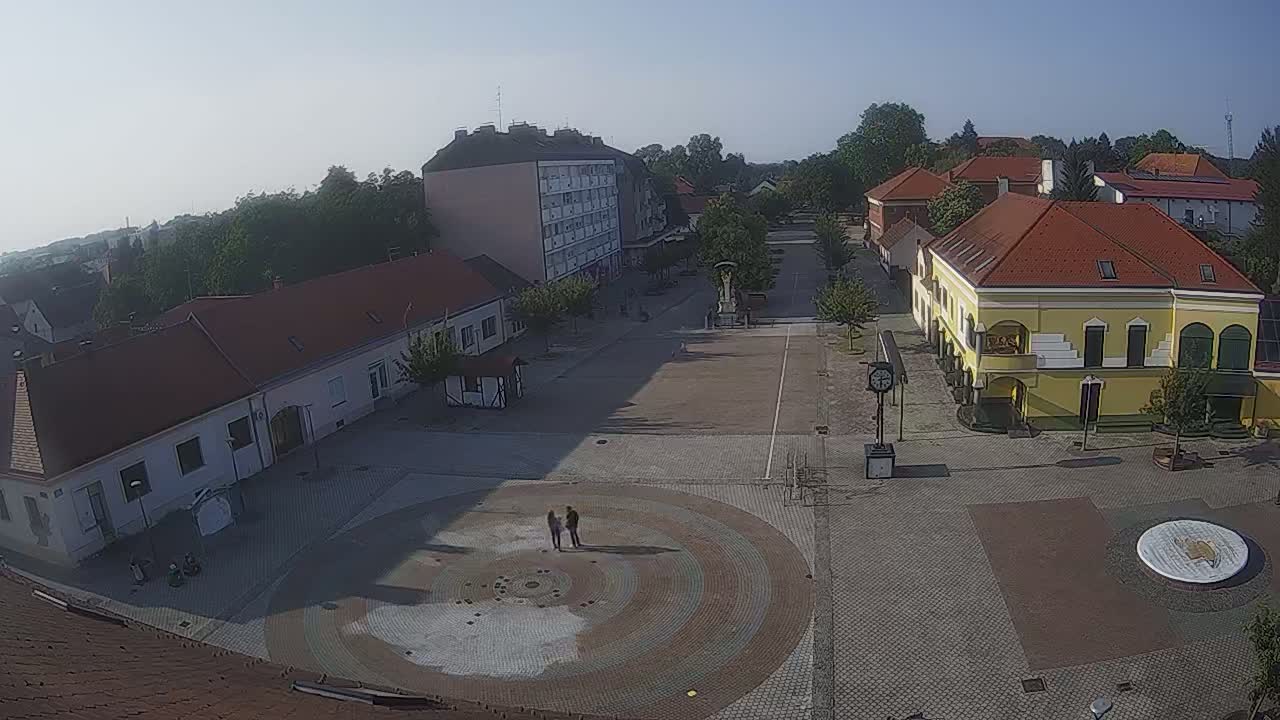A paved town square with several buildings surrounding it, under a clear, sunny sky, features a circular pattern in the center of the square.