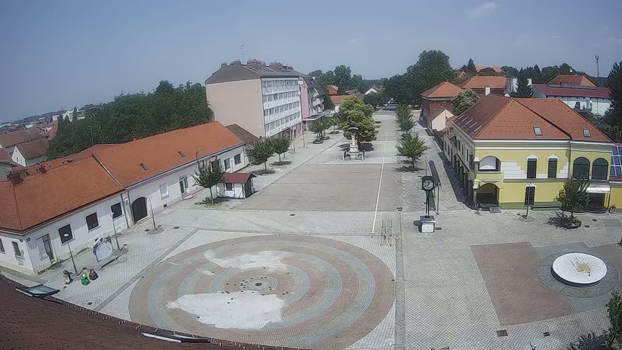 A town square with low buildings and a circular, multicolored paved area is shown on a sunny day.
