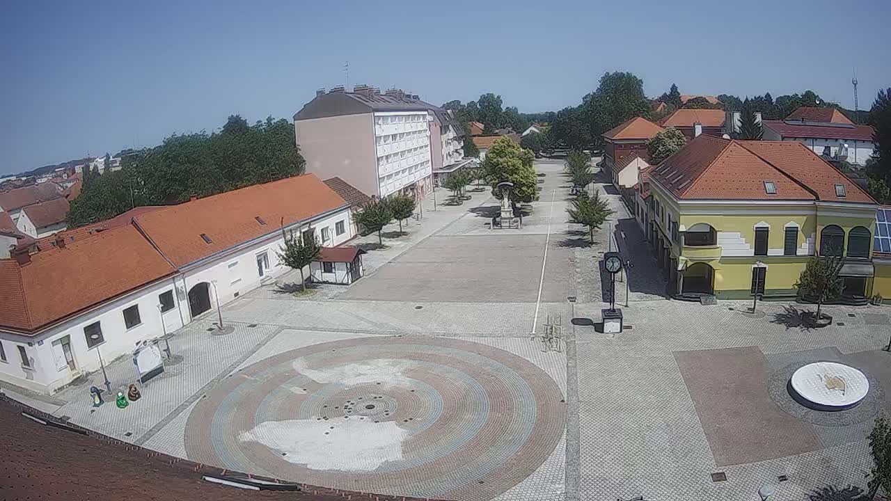 A town square with several buildings and a circular paved area in the center, under sunny skies.