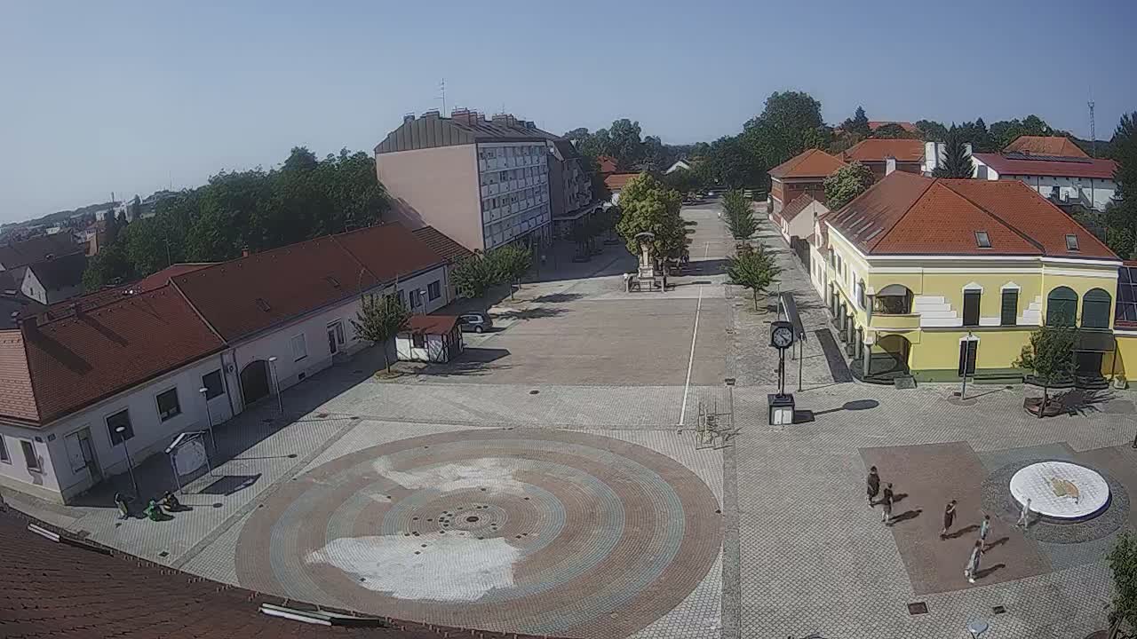 A town square with several buildings surrounding it, a circular paved area in the center, and a few people walking around on a sunny day.