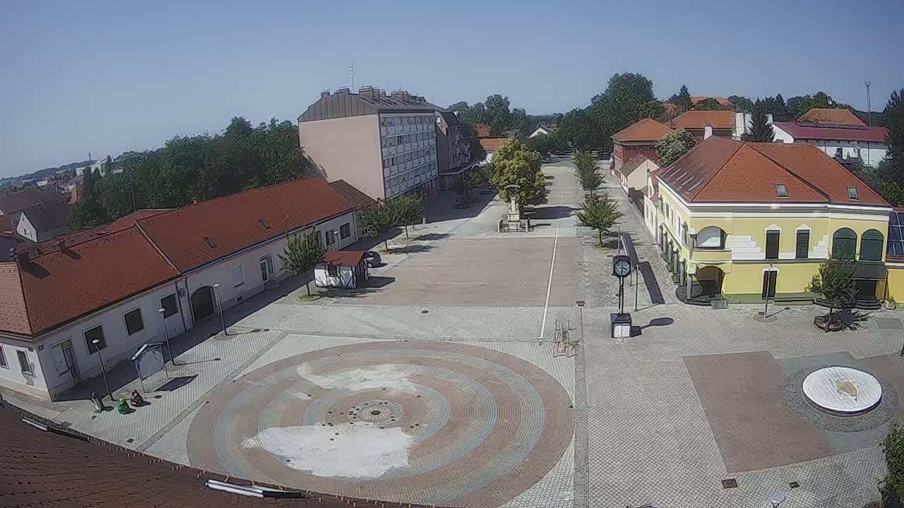 A high-angle view of a town square on a sunny day, showing several buildings with red tile roofs and a large circular paved area in the center.