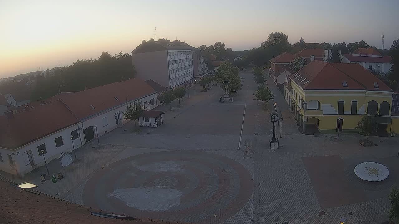 A high-angle view of a town square on a sunny day, showing several buildings with red tile roofs and a large circular paved area in the center.