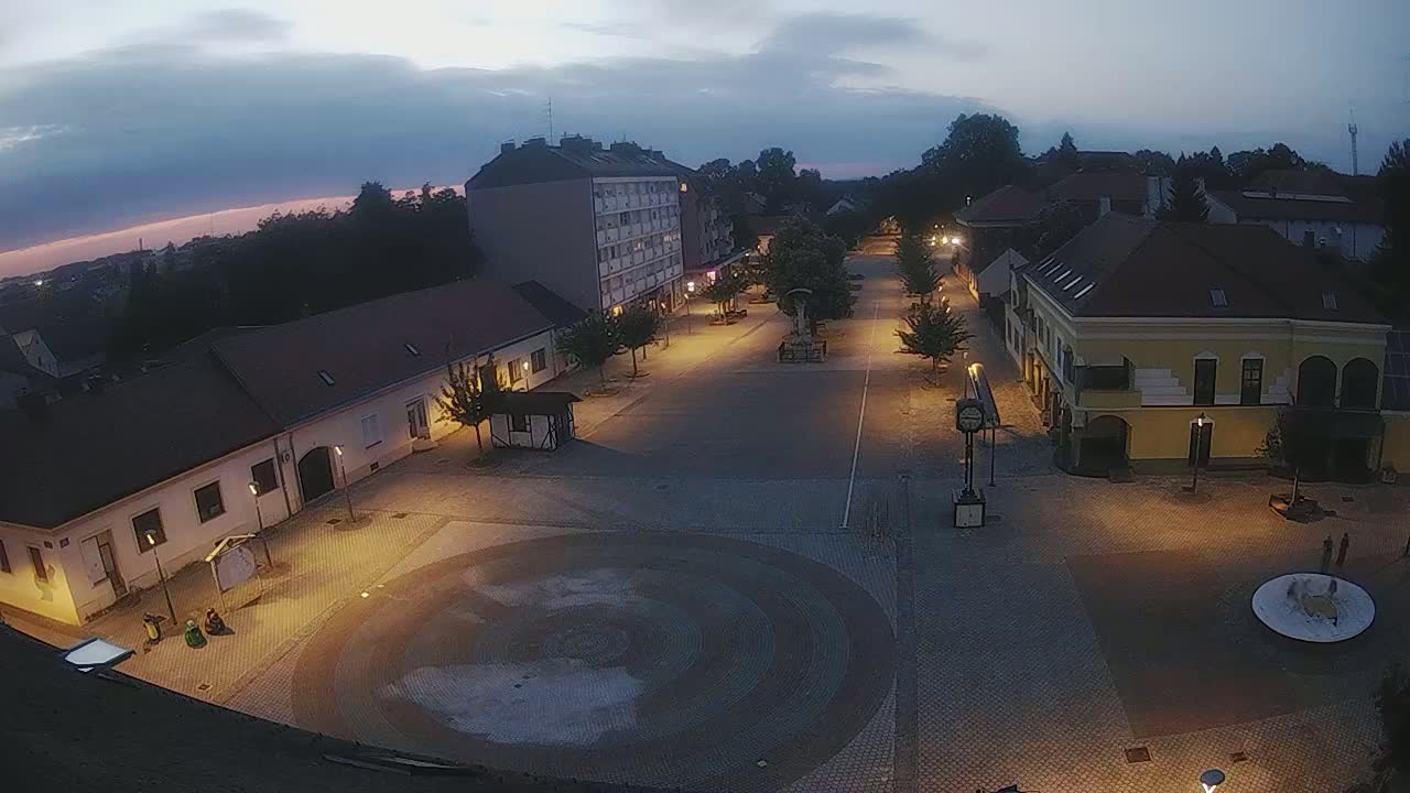 A town square at twilight is illuminated by streetlights, showing buildings lining the sides of a paved area with a circular design in the center.