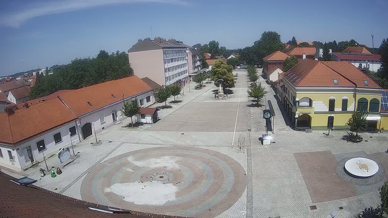 A sunny day reveals a town square with low buildings, trees lining the paved area, and a circular mosaic in the center.