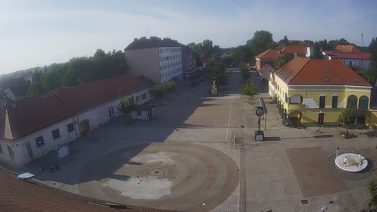 An aerial view of a town square on a sunny day, showing several buildings and a circular paved area in the center.
