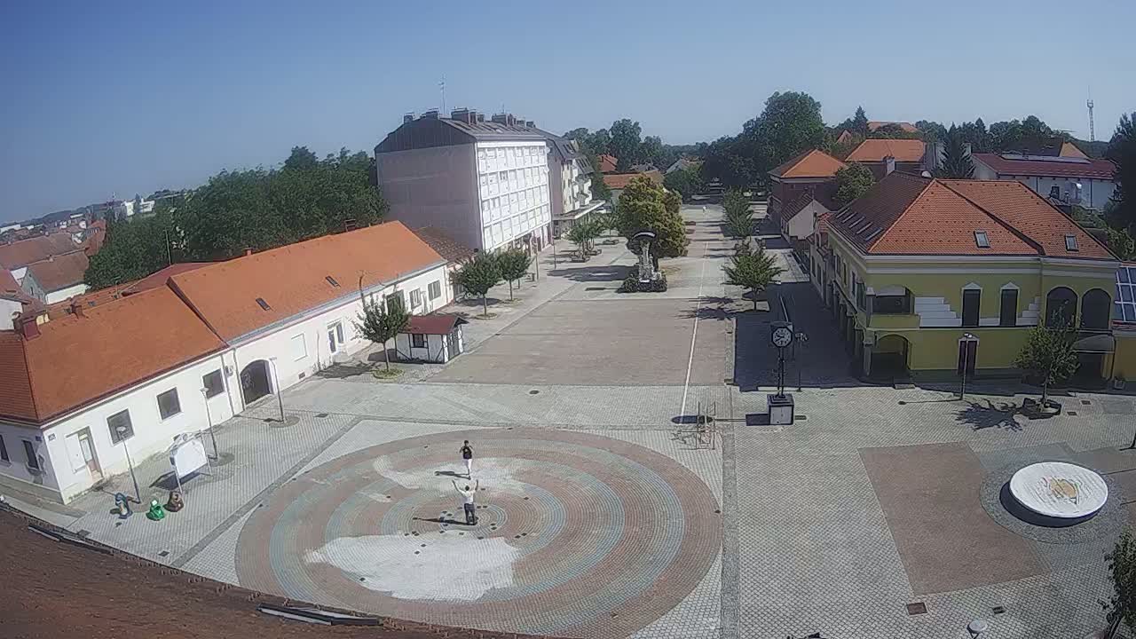 A town square with several buildings, a circular paved area, and a few people, under a clear, sunny sky.