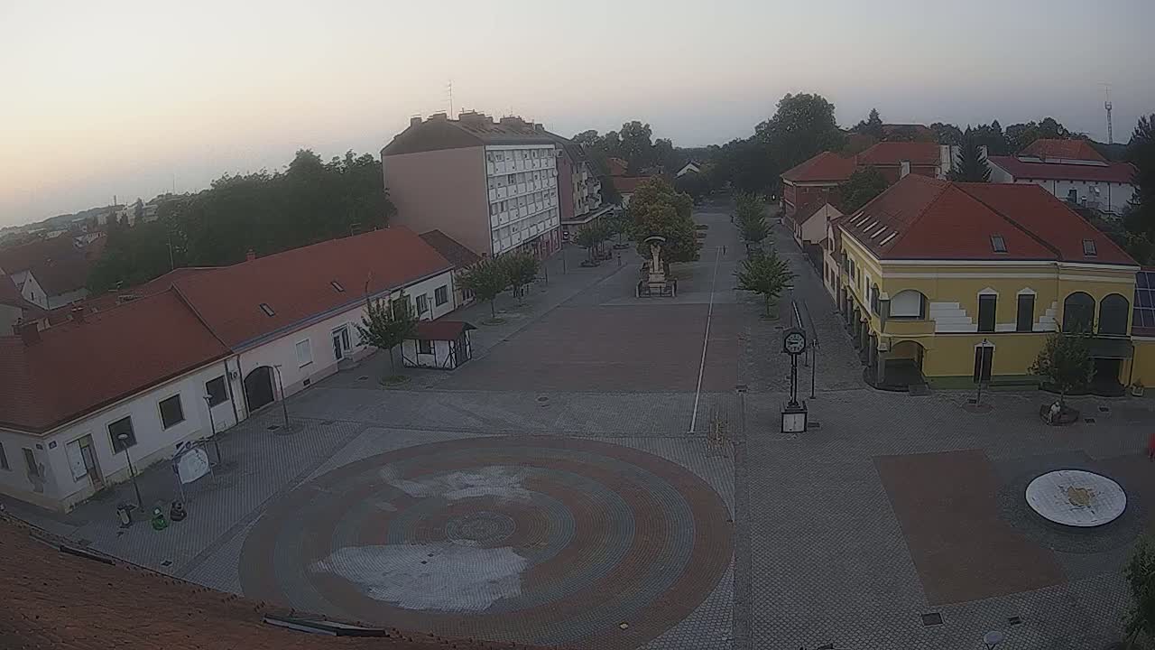 A town square with low-rise buildings, a central paved area featuring a circular mosaic, and trees, under an overcast sky.