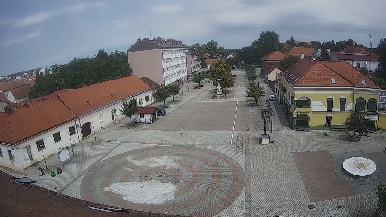 An overcast day reveals a town square with low buildings, a central paved area featuring a circular design, and sparsely parked cars.