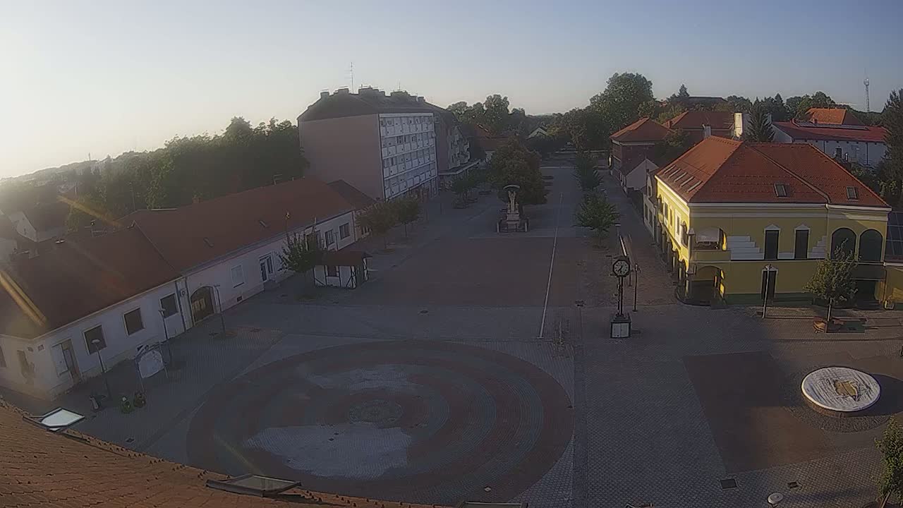A town square with several buildings, trees, and a circular paved area is visible under sunny, clear skies.
