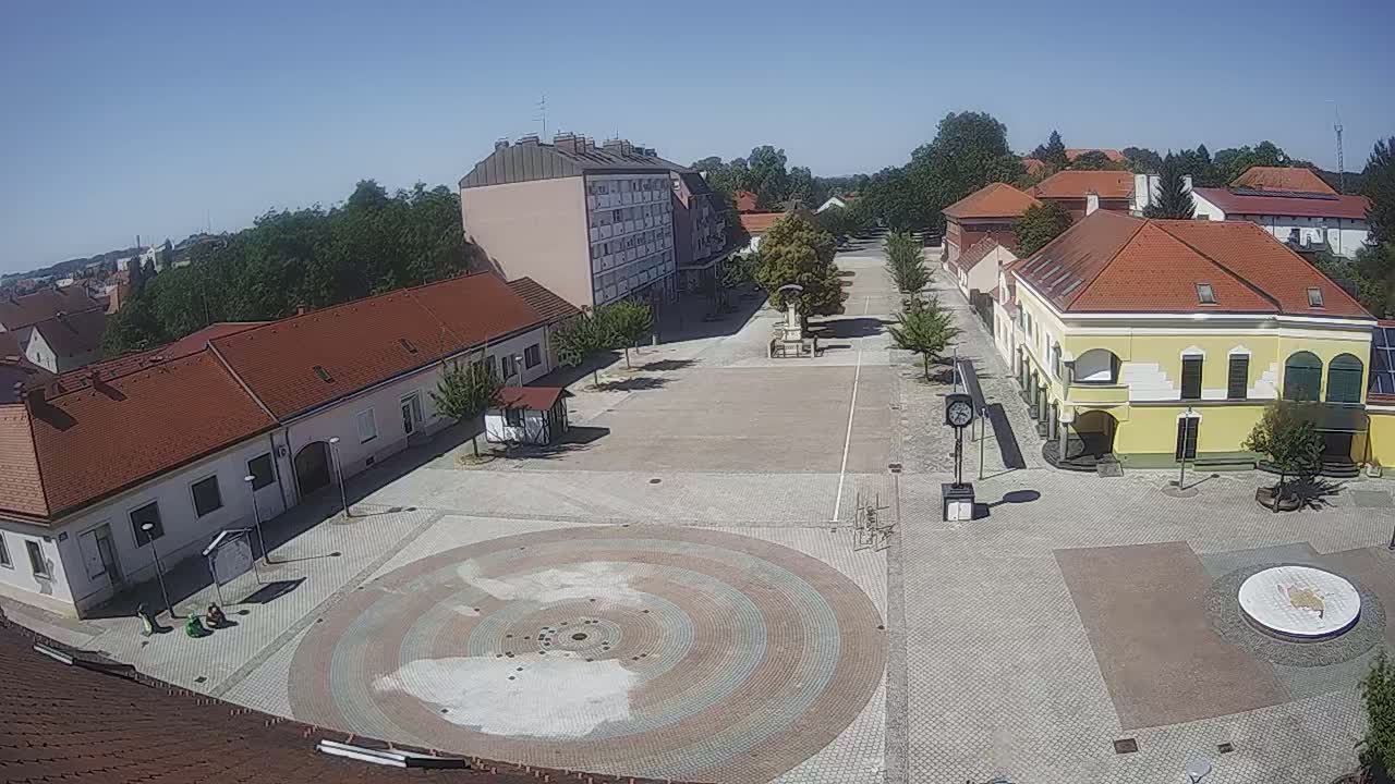 A town square with several buildings and a circular paved area in the center, under sunny skies.