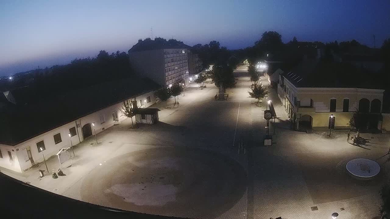 A mostly empty town square at night, illuminated by streetlights, is surrounded by low buildings under a twilight sky.