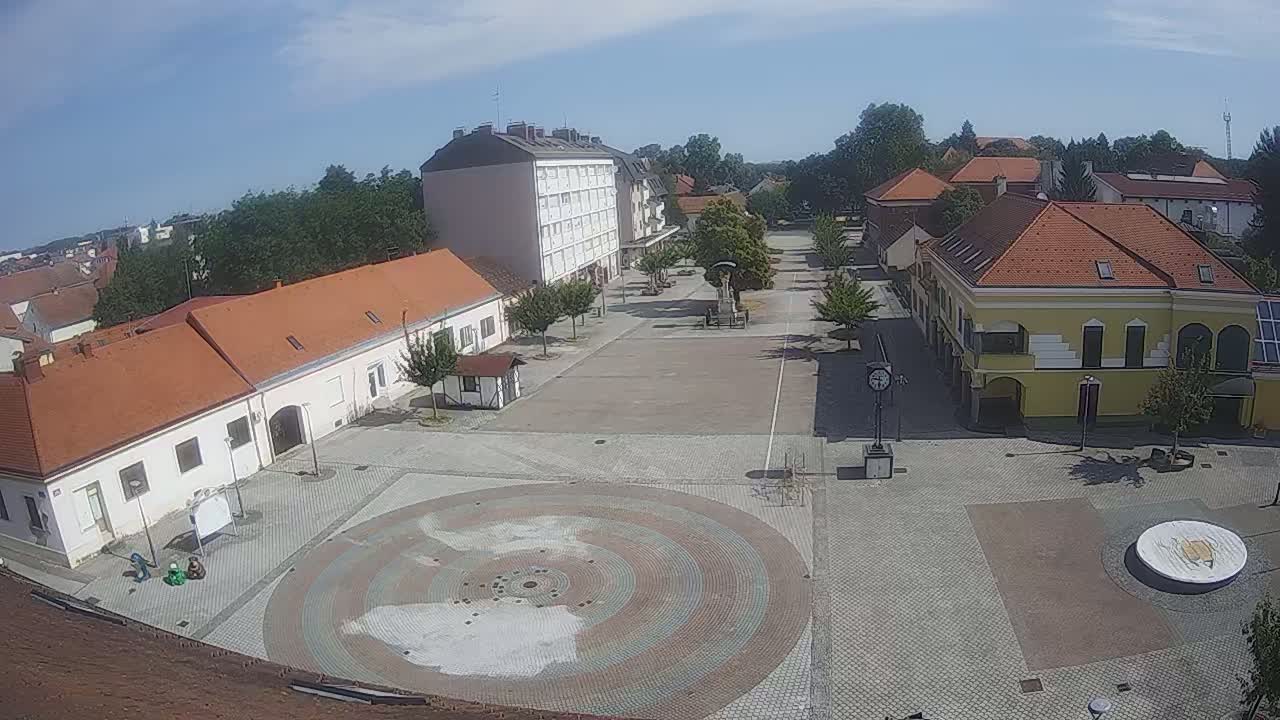 A town square with several buildings surrounding it, under sunny skies, features a circular paved area in the center.