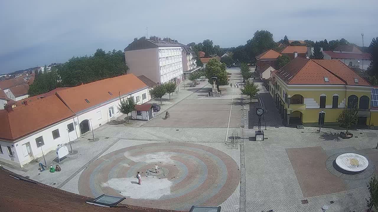 A town square with several buildings and a circular paved area in the center, under sunny skies.