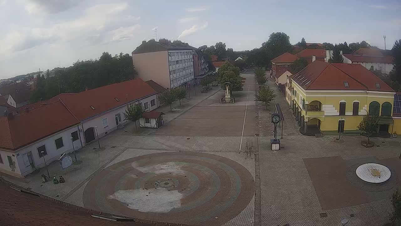 A town square with several buildings and a circular paved area in the center, under sunny skies.
