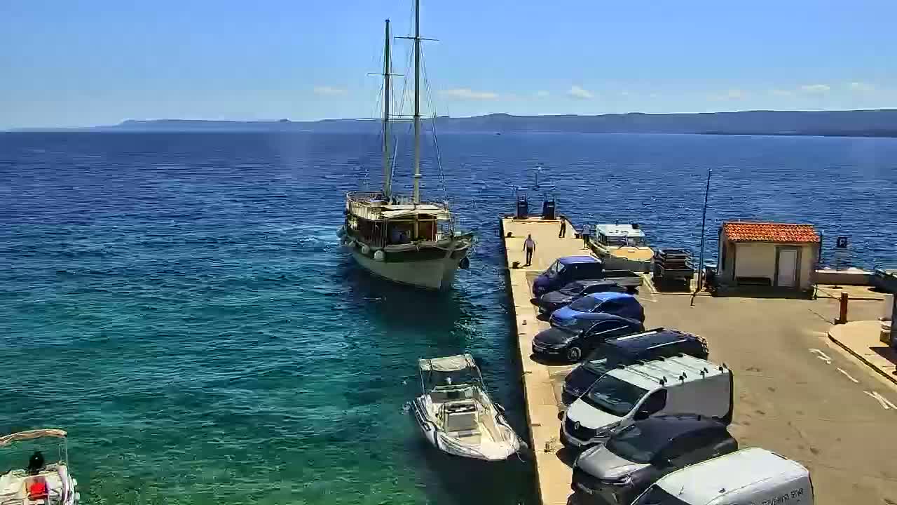 A sailboat is docked at a pier next to a parking lot filled with cars and vans on a sunny day with clear blue water.