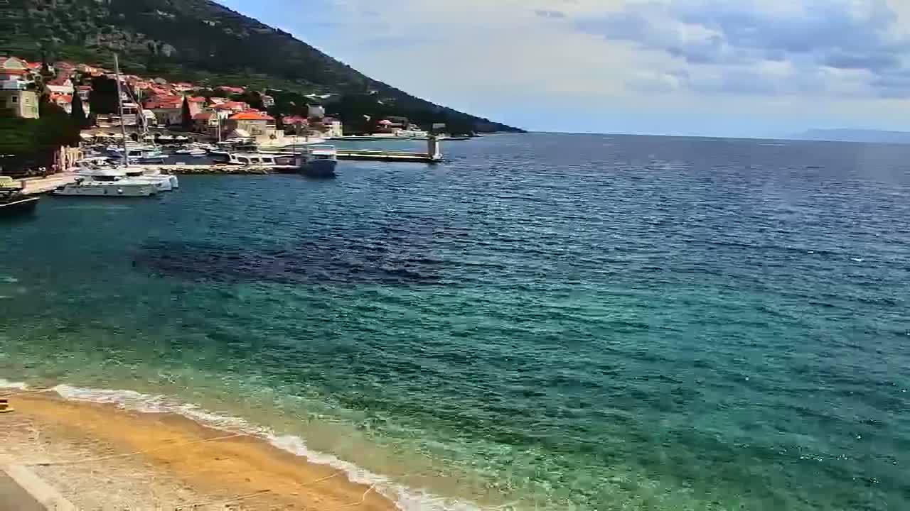 A small town nestled against a hillside with a harbor filled with boats sits on a calm, clear sea with a sandy beach in the foreground under a partly cloudy sky.