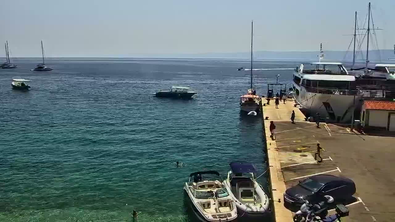 A sunny day at a harbor shows several boats docked and in the water, with people walking along a pier.