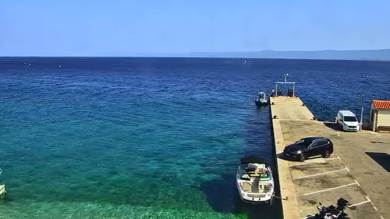 A long pier extends into clear, blue-green water on a sunny day, with several boats moored nearby and a few vehicles parked on the pier's landside.