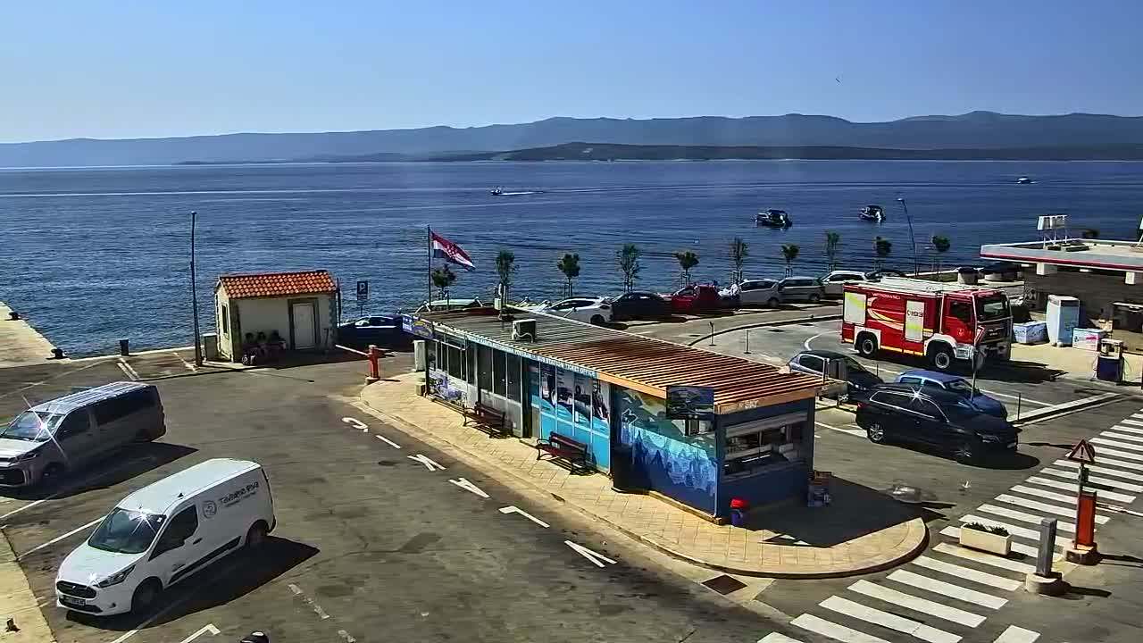 A harbor scene on a sunny day shows a parking lot with several cars and vans near a small kiosk and a firetruck, overlooking a calm sea with distant mountains and a few boats.