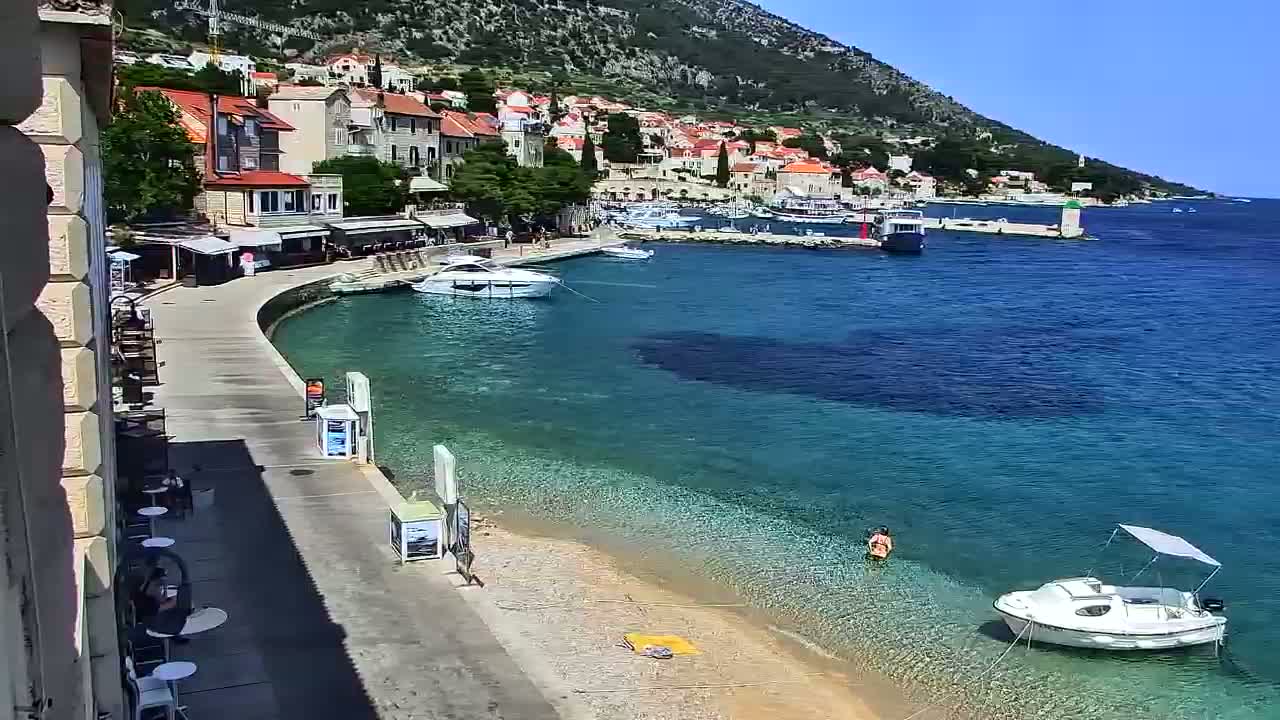 A sunny day at a seaside town shows a small beach with clear, shallow turquoise water, several boats, and buildings along a curved waterfront.