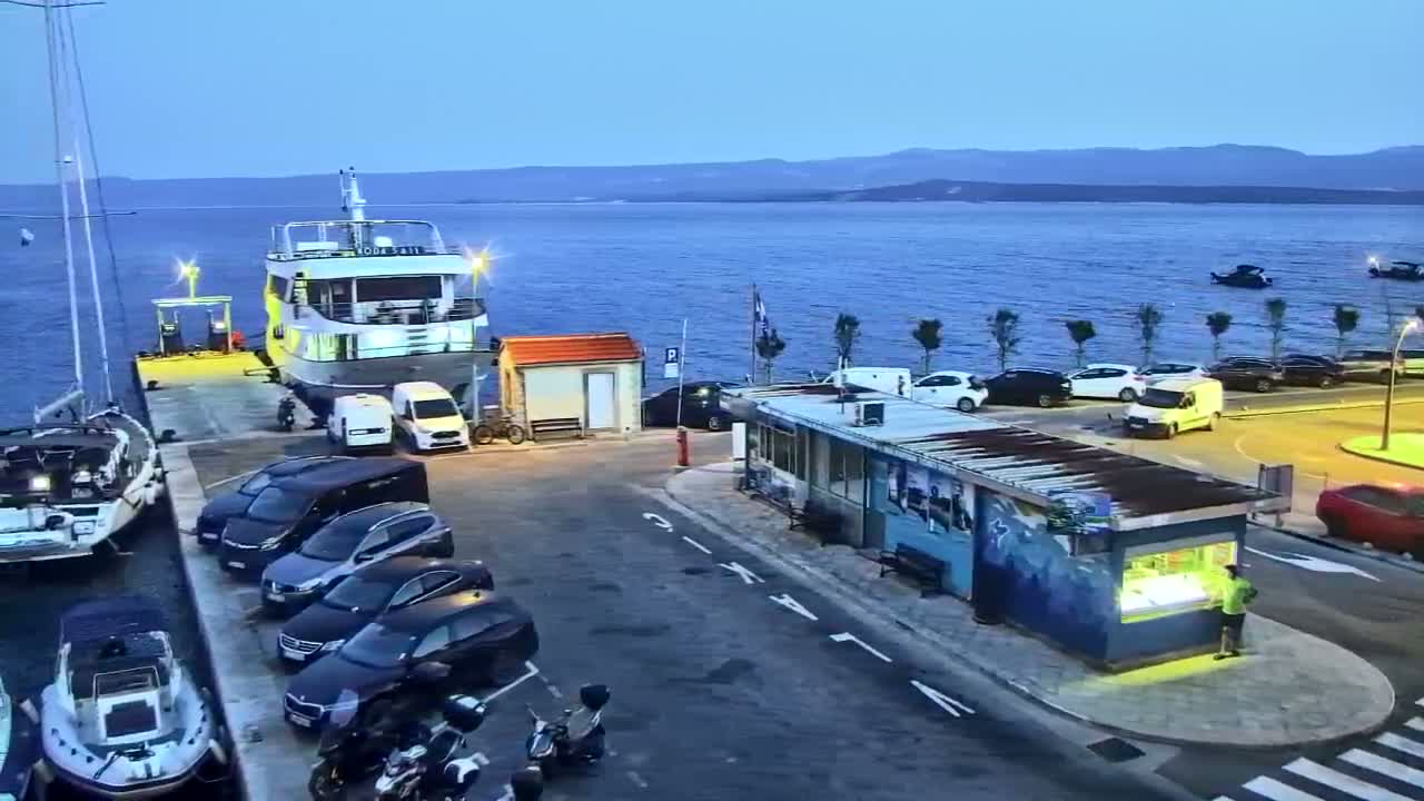 A sunny day at a seaside town shows a small beach with clear, shallow turquoise water, several boats, and buildings along a curved waterfront.