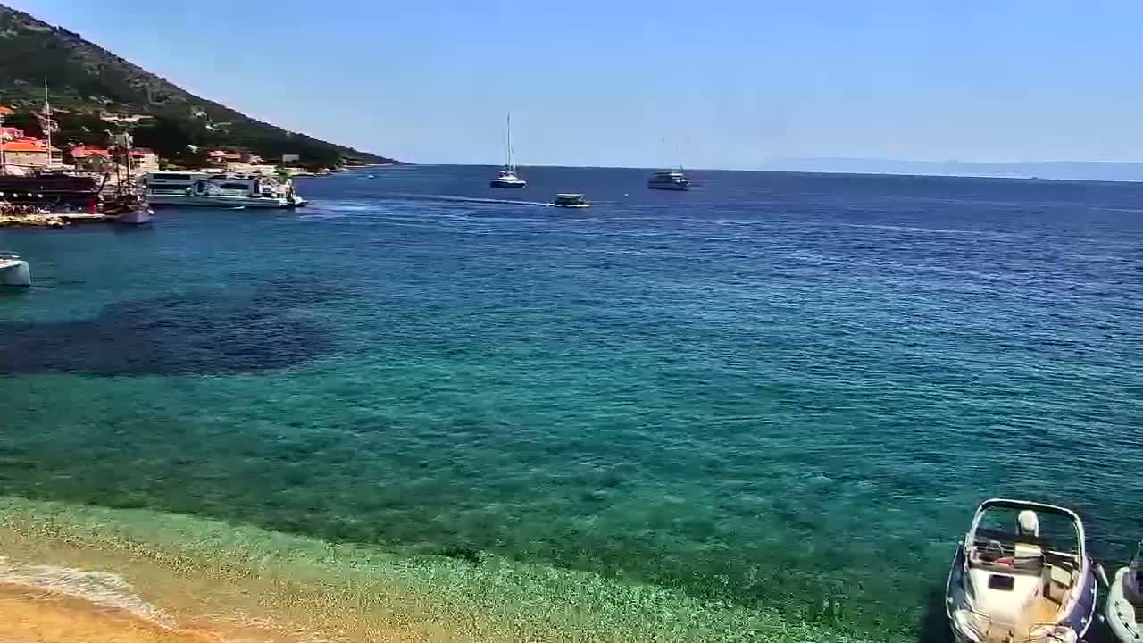 A clear, sunny day reveals a calm, turquoise sea with several boats docked near a shoreline with buildings and a small sandy beach.