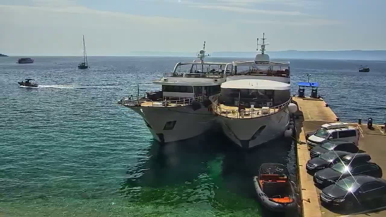 Two large yachts are docked at a pier on a sunny day, with several cars parked nearby and smaller boats visible in the clear blue water.