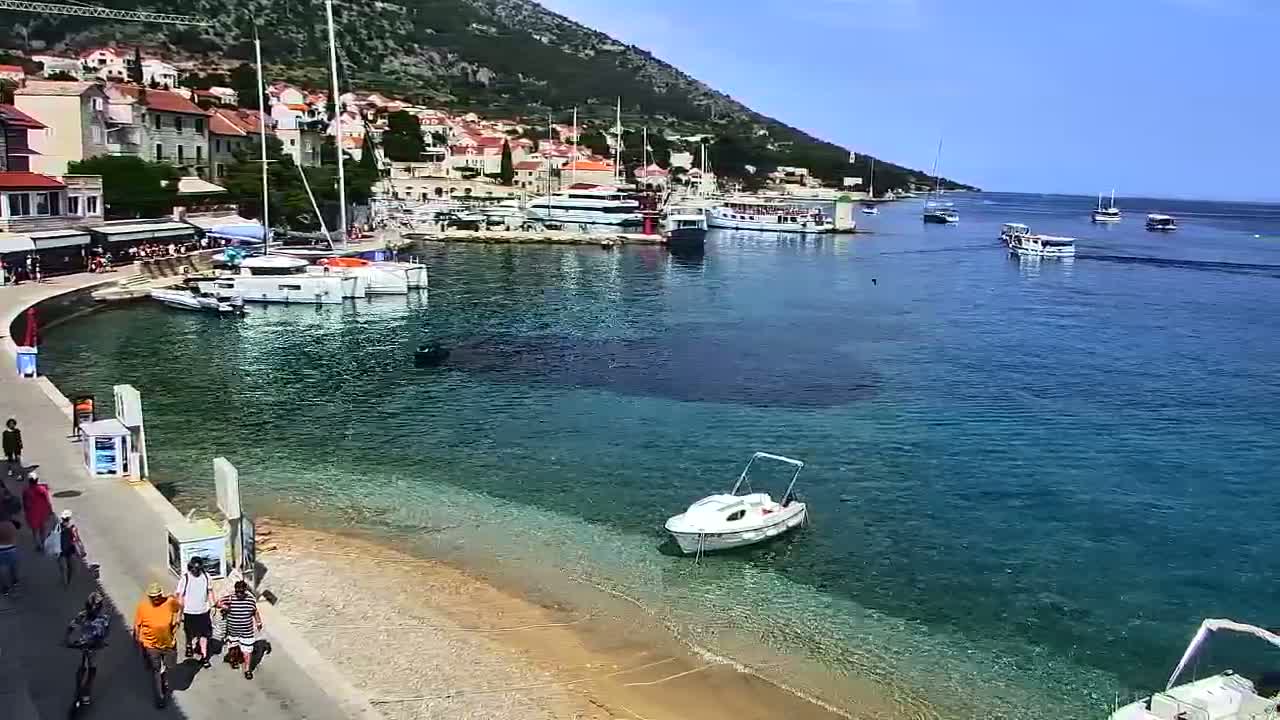 A picturesque harbor town nestled against a hillside, under sunny skies, features numerous boats in clear, calm water, with people strolling along a nearby walkway.