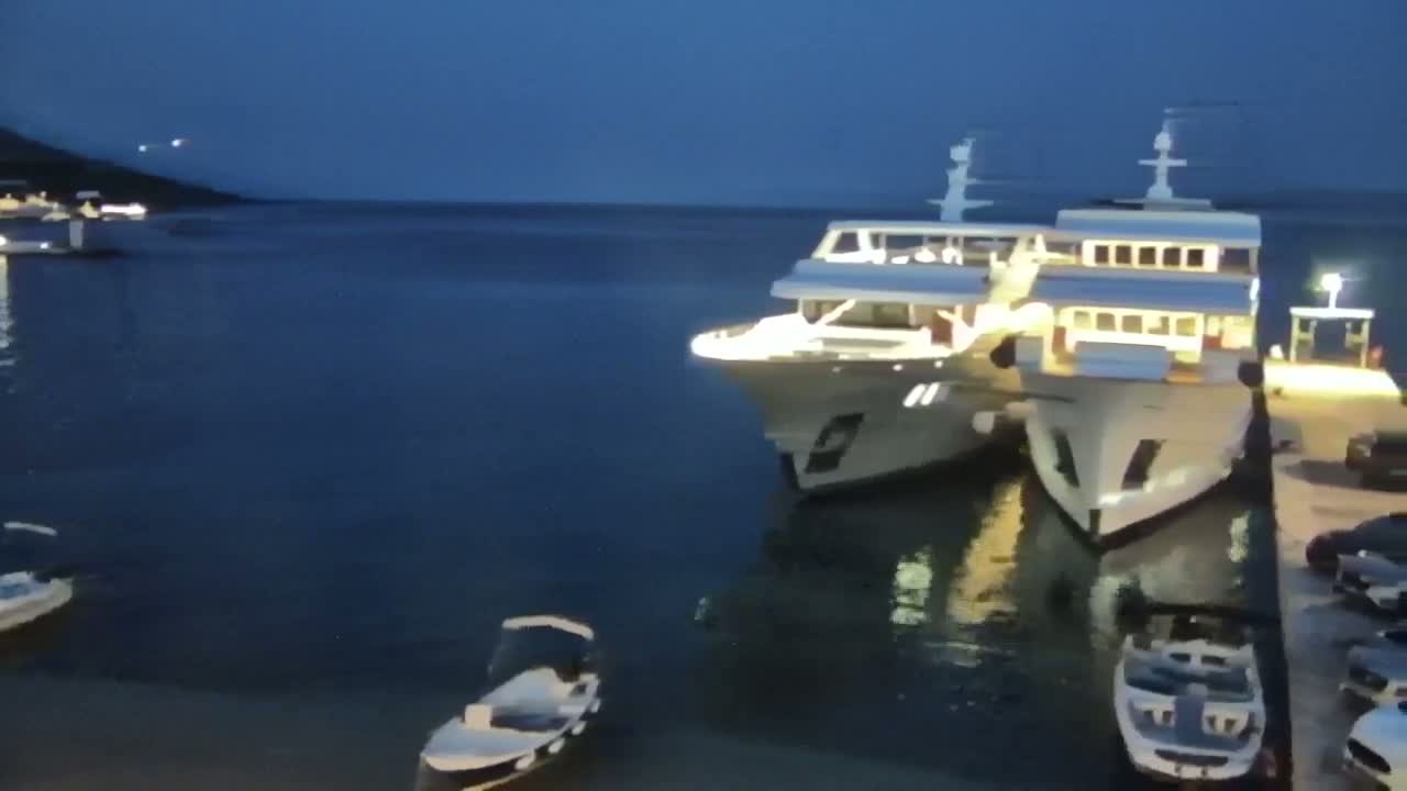 Two large yachts are docked at a harbor at night, with smaller boats visible in the water and cars parked nearby under a dark sky.
