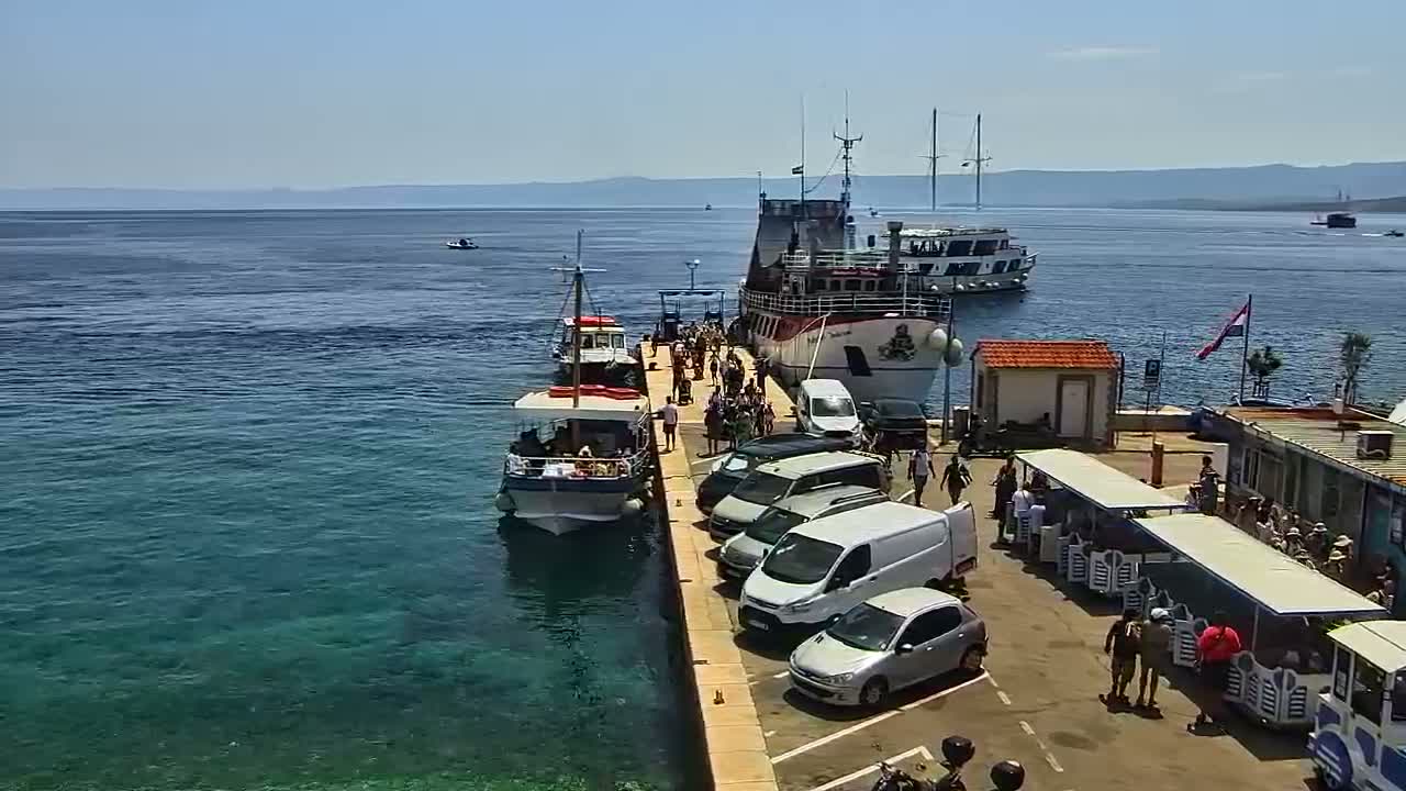 A ferry and several smaller boats are docked at a pier on a sunny day, with numerous people disembarking and cars parked nearby.