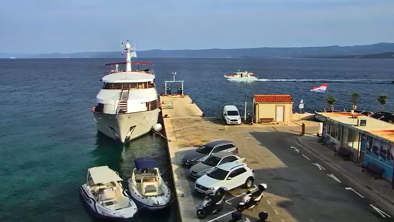 A large yacht is docked at a marina on a sunny day, with several cars and smaller boats nearby and another boat in the distance.