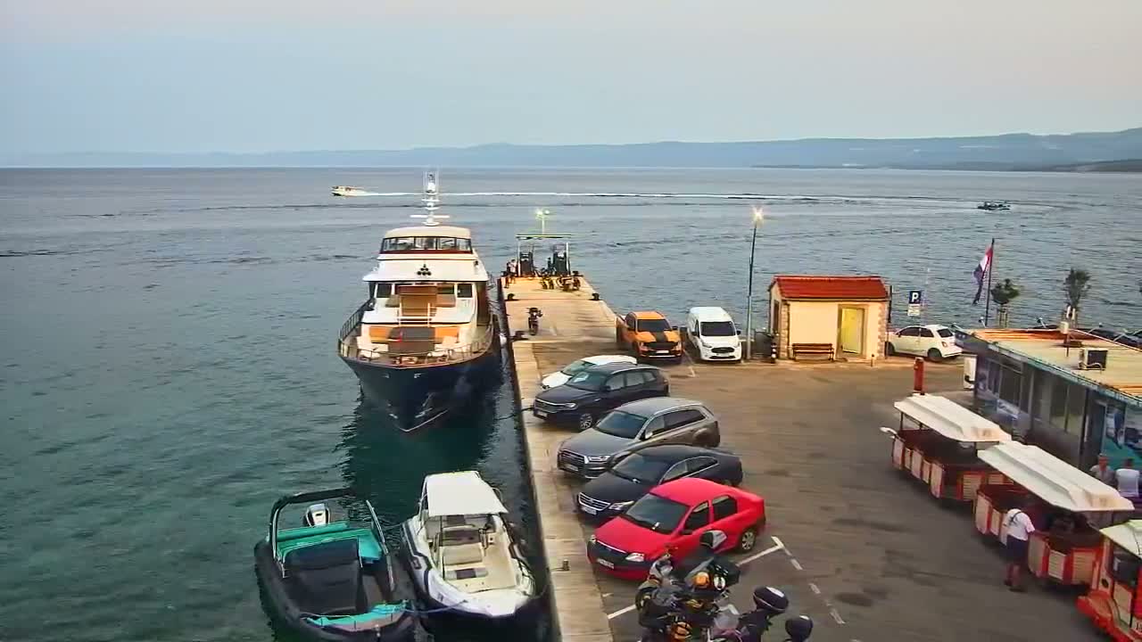 A large yacht is docked at a pier next to a parking lot with several cars and small boats, all under a cloudy sky overlooking a calm sea.