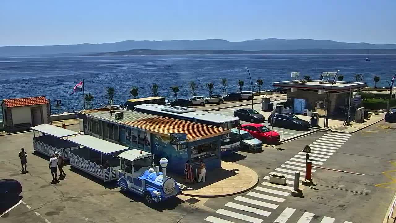 A seaside parking lot with a small train, buses, cars, and a kiosk, on a sunny day with clear blue water and distant mountains in the background.