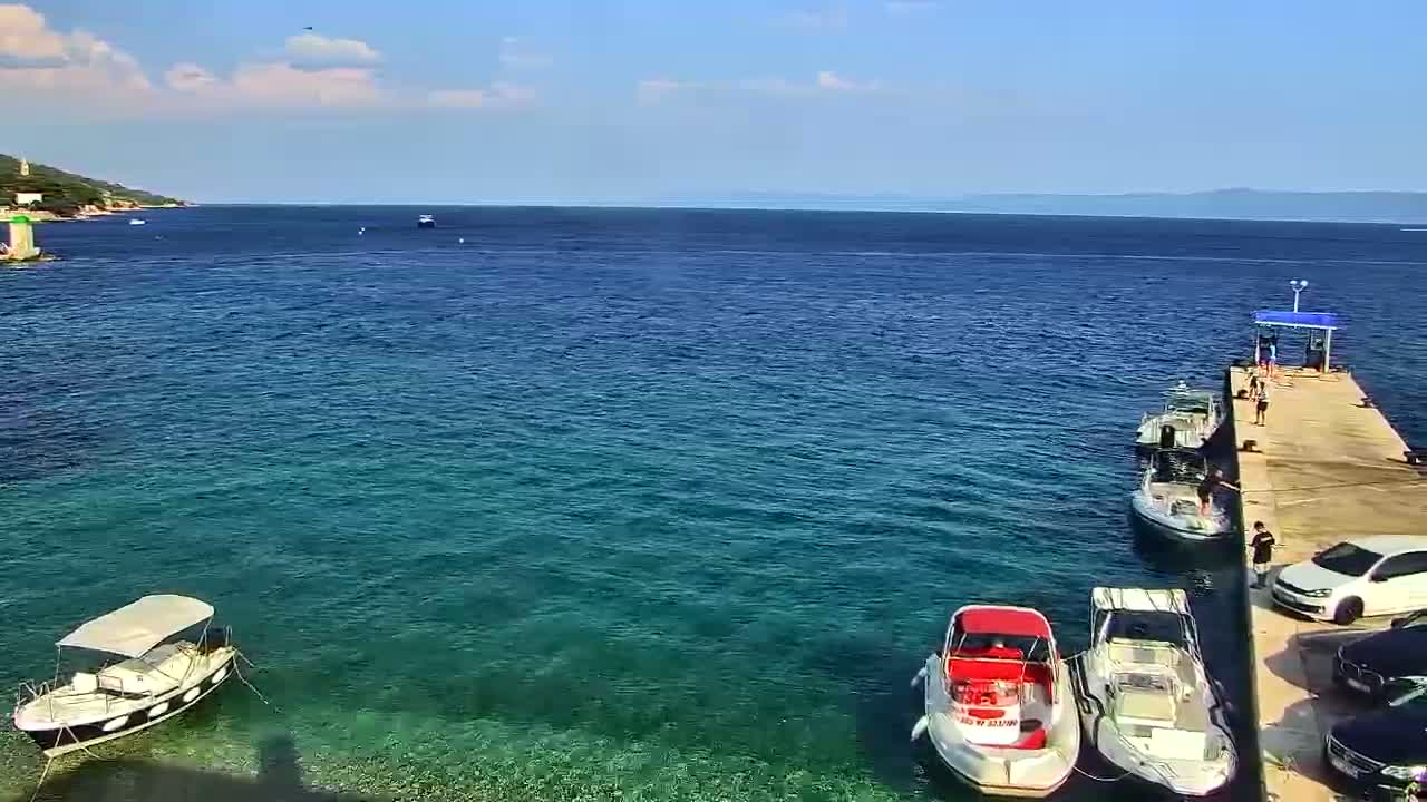 A clear, sunny day shows a calm, teal-colored sea with several boats moored at a pier, alongside a shoreline with some vegetation and a small building.