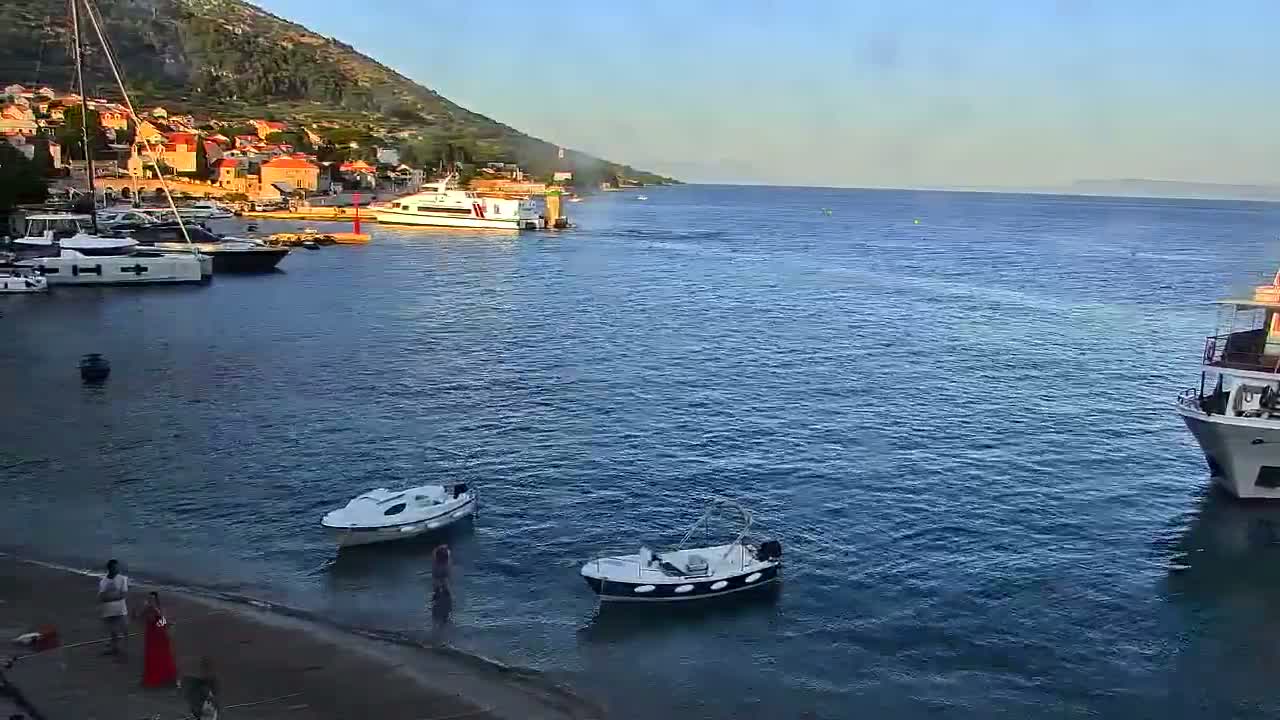 A calm, clear day reveals a harbor filled with boats near a hillside town, with additional boats and a ferry visible further out in the water.