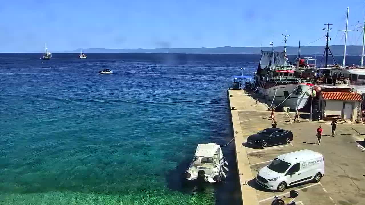 A sunny day shows a harbor with clear blue water, several boats, and a few cars parked near a docked ship.