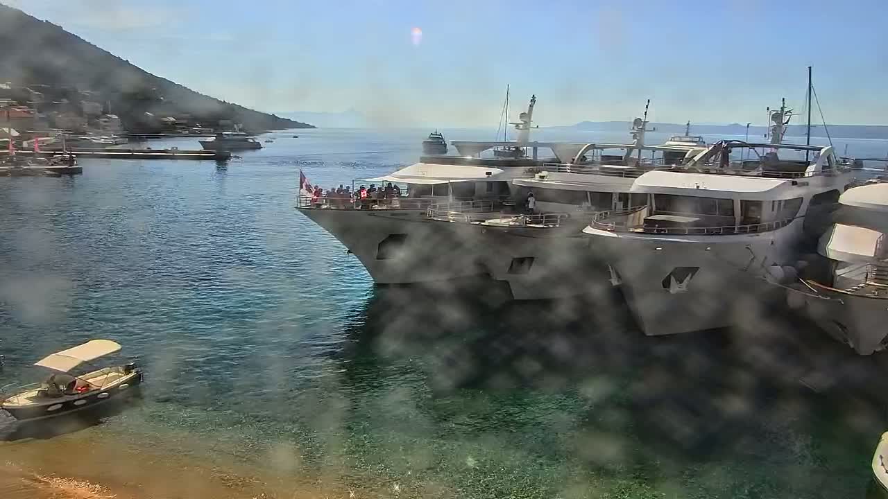 Several large yachts are docked in a calm harbor near a hillside town on a sunny day.