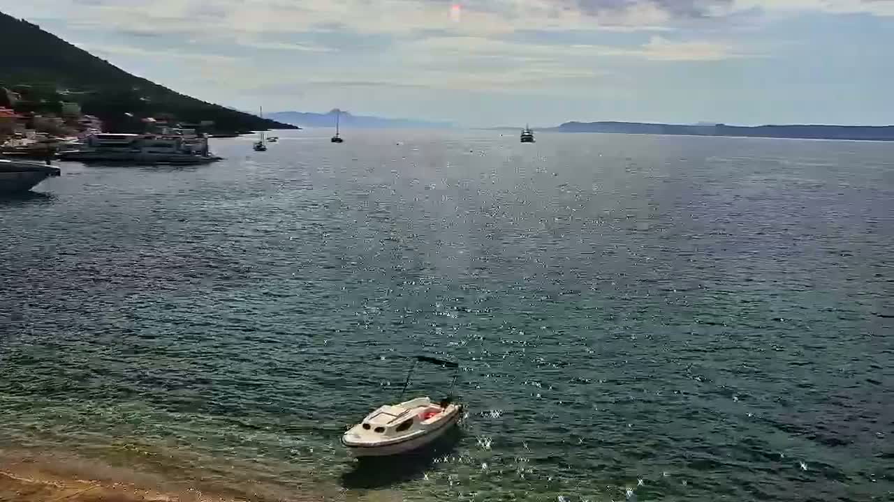 A small white boat sits in calm, clear water near a shoreline with several other boats and a mountainous background under a partly cloudy sky.