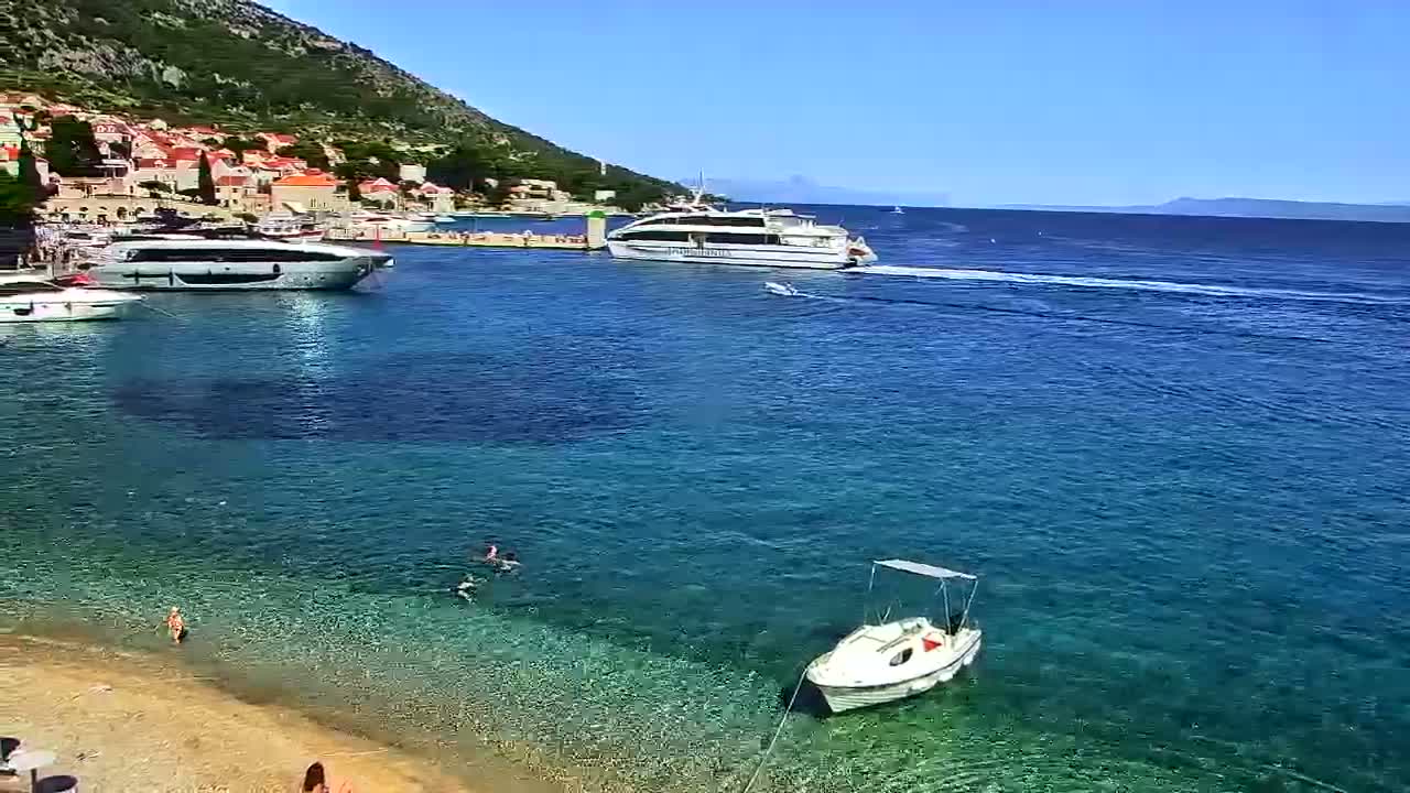 A small white boat sits in calm, clear water near a shoreline with several other boats and a mountainous background under a partly cloudy sky.