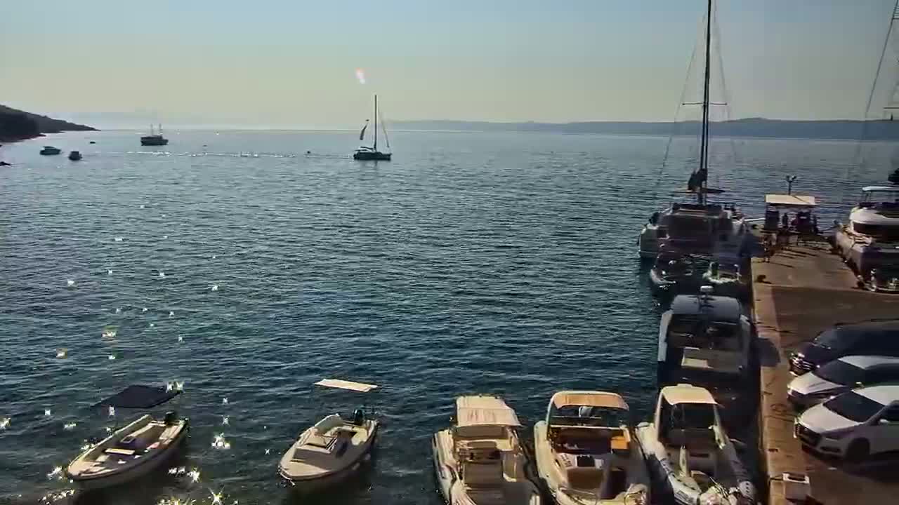 Several boats are moored at a dock on a calm, sunny day, with additional boats and sailboats visible further out on the water.