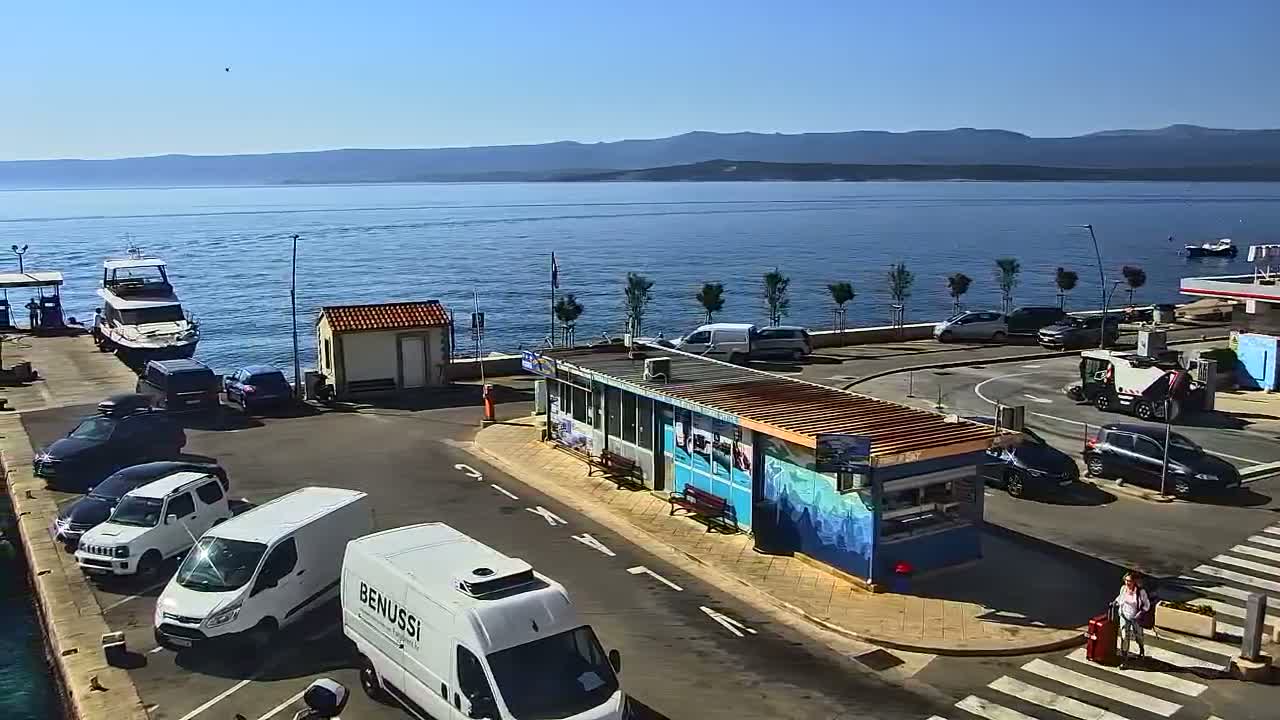 A harbor scene on a sunny day shows several vehicles parked near a small building by the water, with boats moored and mountains visible in the distance.