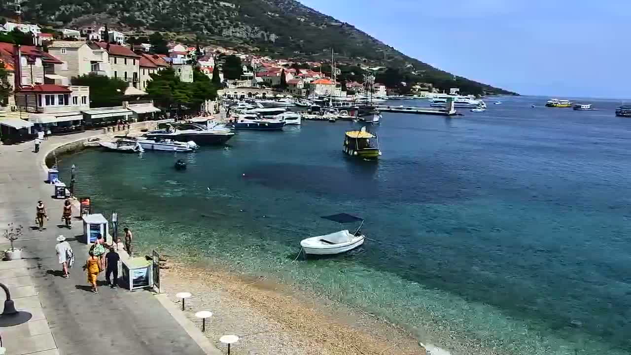 A harbor scene on a sunny day shows several vehicles parked near a small building by the water, with boats moored and mountains visible in the distance.