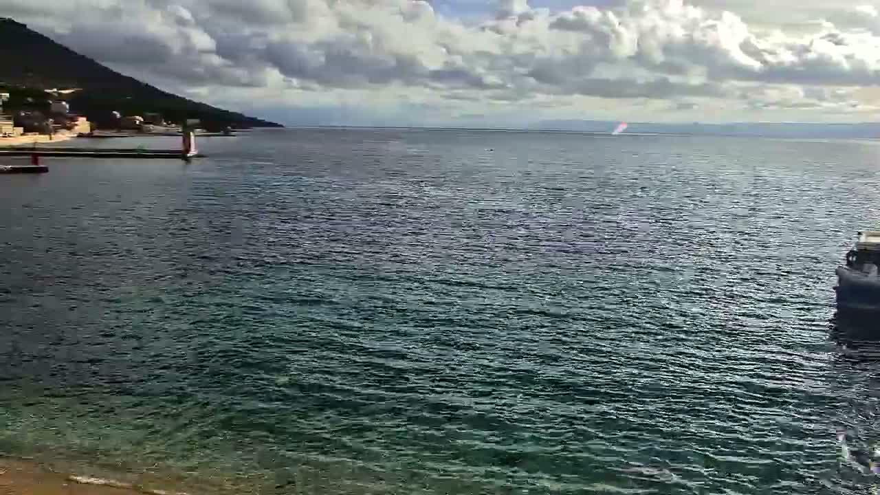 A partly cloudy daytime view shows a calm sea with a mountainous coast and small port to the left, a boat in the right foreground, and a distant paraglider or boat with a wake under a sky dotted with white and grey clouds.