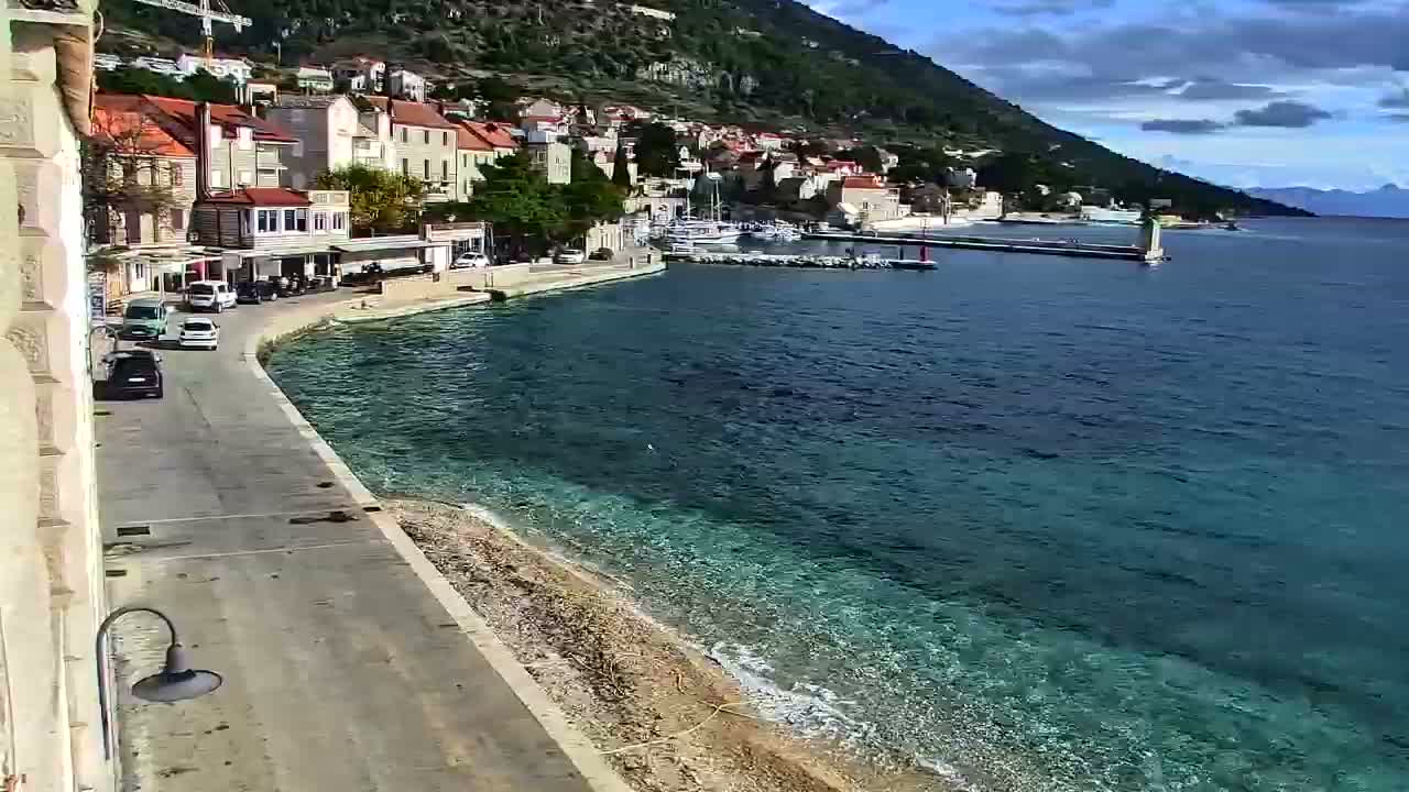 A coastal town with red-roofed buildings clings to a green mountainside overlooking a clear turquoise bay with a small harbor and a curving road, all bathed in bright daylight under a partly cloudy sky.