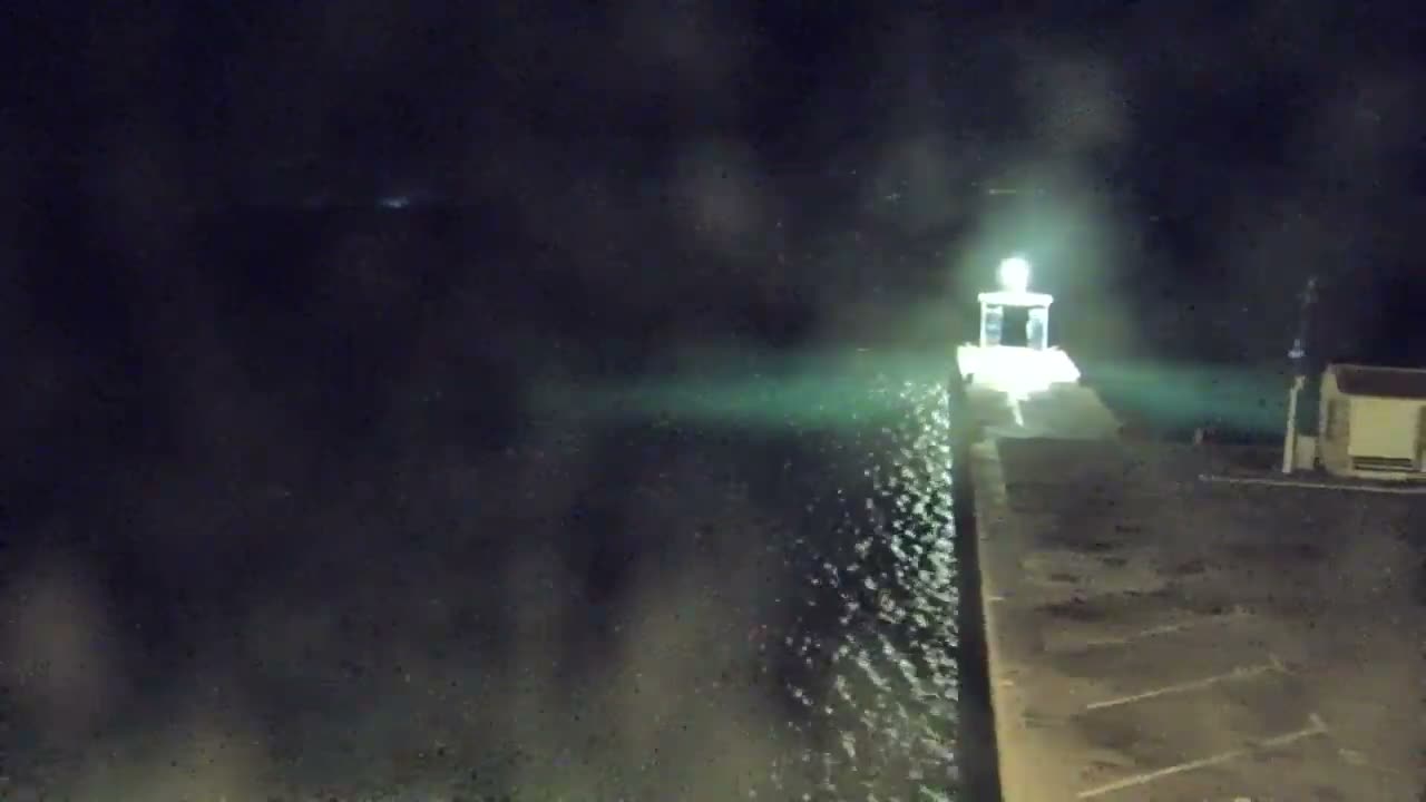 A brightly lit beacon at the end of a pier shines over dark, rippling water and a wet paved area on a rainy night, with faint lights visible in the distant mist.