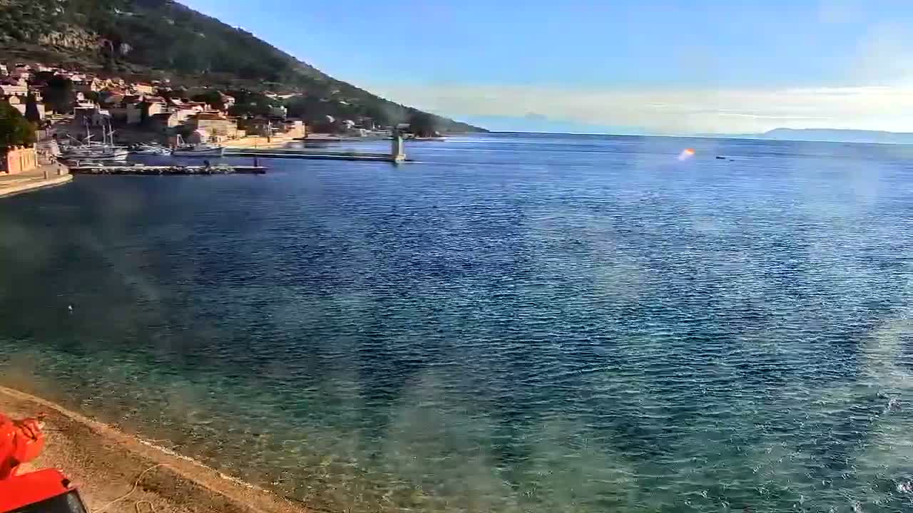 On a clear, sunny day, a picturesque coastal town with houses built into a green hillside overlooks a calm bay featuring moored boats and a lighthouse on a pier, with clear blue-green waters stretching to the horizon where a bright orange light or reflection is visible.