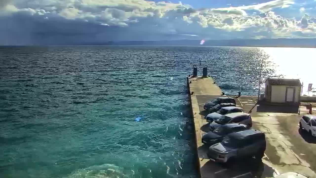 A concrete pier lined with parked cars extends into a vast expanse of turquoise water under a sky marked by a mix of bright sunshine, white fluffy clouds, and darker, heavier storm clouds, with distant land visible on the horizon.